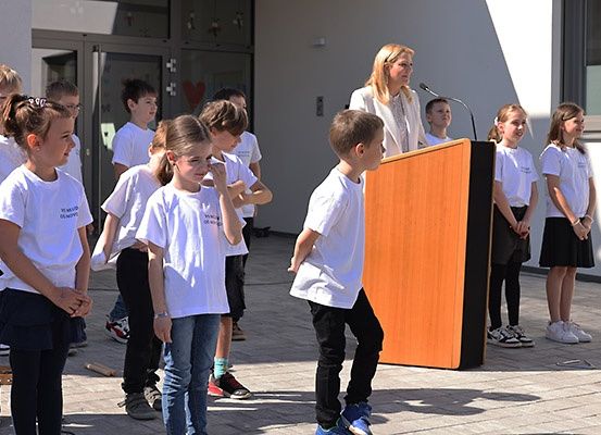 Eine Gruppe von Kindern in weißen T-Shirts ist um ein Podium versammelt, an dem eine Frau bei einer Veranstaltung im Freien spricht.