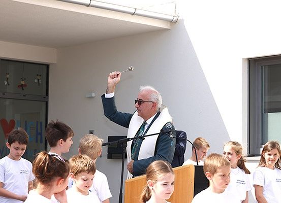 Ein Mann in einer weißen Robe hebt eine Löffel am Rednerpult vor einer Gruppe von Kindern in weißen Hemden.