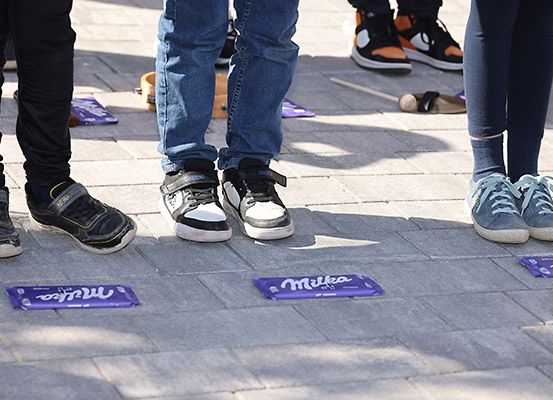 Eine Gruppe von Personen steht auf einem Ziegelboden mit verschiedenen Gegenständen, darunter Schuhe, ein Korb und Schokoladentafeln. Milka-Schokoladentafeln sind auf dem Boden sichtbar.