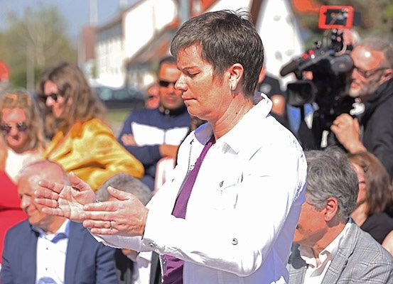 Eine Frau in einem weißen Hemd und einer lila Krawatte steht vor einer Menschenmenge und scheint zu sprechen. Hinter ihr hält ein Mann in einem Anzug eine Kamera. Die Menschen im Hintergrund tragen verschiedene Kleidungsstücke.