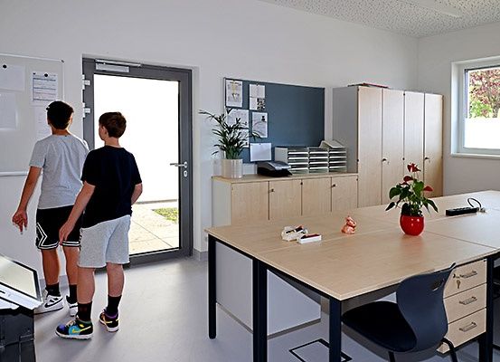 Zwei Jungen stehen in einem Klassenzimmer in der Nähe einer Tür und sehen sich etwas auf einem Tisch mit Fernbedienung und Topfpflanze an.