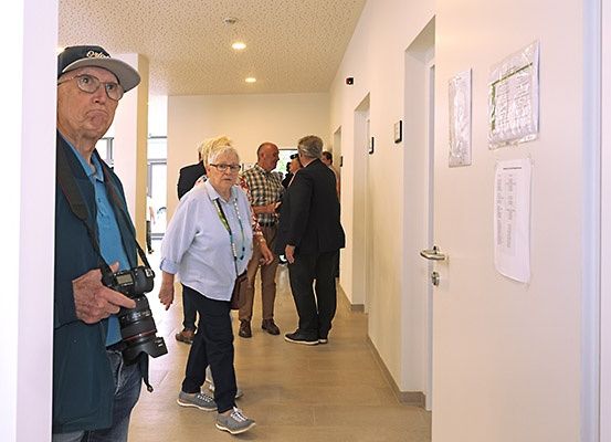 Eine Gruppe von Menschen, einschließlich eines älteren Mannes mit einer Kamera, geht in einem Flur. Papiere hängen an der Wand.