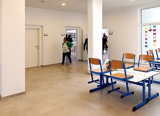 Ein leerer Klassenraum mit weißen Wänden, blauen Tischen und Stühlen. Eine Frau geht durch den Flur.