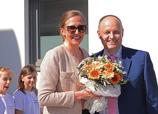 Ein Mann und eine Frau stehen lächelnd nebeneinander und halten einen Blumenstrauß. Sie sind formell gekleidet, die Frau trägt eine Sonnenbrille. Hinter ihnen stehen zwei junge Mädchen in weißen Hemden und lächeln ebenfalls.