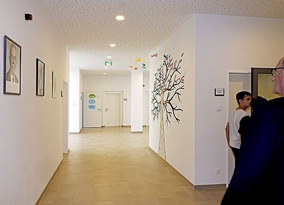 Ein Flur mit gefliesten Böden, weißen Wänden und einer Wandmalerei eines Baumes. Es gibt gerahmte Bilder auf der linken Seite und ein Mann, der in einen Raum auf der rechten Seite schaut.