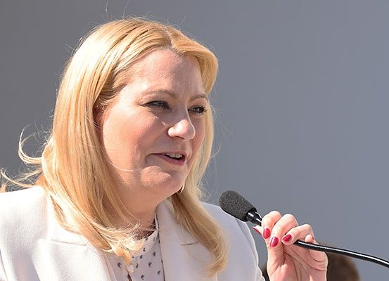 Eine Frau mit blonden Haaren spricht in ein Mikrofon, während sie einen weißen Blazer und ein gepunktetes Hemd trägt.