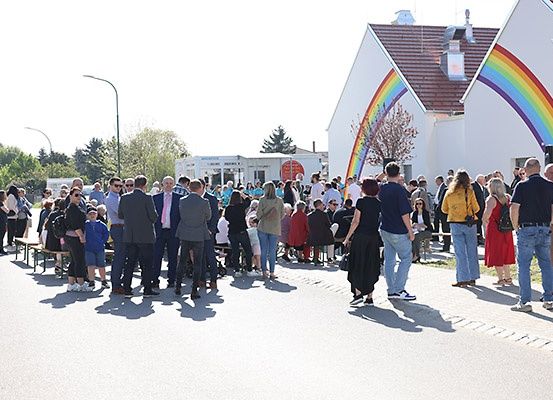 Eine Menschenmenge steht draußen, einige mit Masken, in der Nähe eines Gebäudes mit einem Regenbogenbogen. Eine Bank ist in der Nähe.