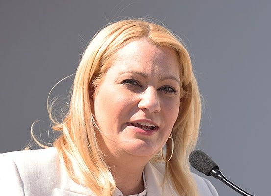 Eine Frau mit blonden Haaren steht lächelnd vor einem Mikrofon. Sie trägt einen weißen Mantel.