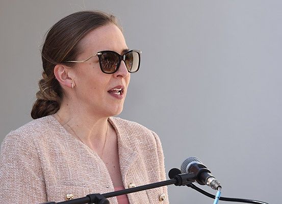 Eine Frau mit Sonnenbrille steht an einem Rednerpult mit einem Mikrofon, trägt ein rosa Hemd und einen beigen Blazer. Sie hält wahrscheinlich eine Rede.