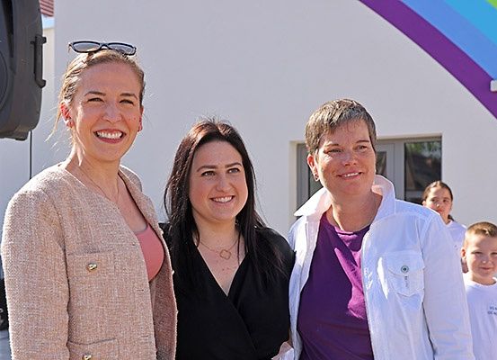 Drei Frauen lächeln für die Kamera vor einem Gebäude mit einem Regenbogen an der Wand. Ein Lautsprecher und einige Kinder sind ebenfalls zu sehen.