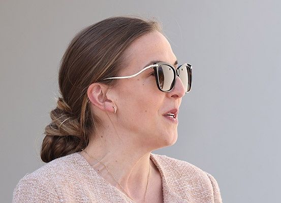 Eine Frau mit Duttfrisur und Silbersonnenbrille schaut nach rechts. Sie trägt einen rosa Blazer und eine Halskette.
