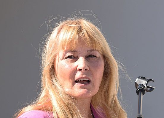 Eine Frau mit blonden Haaren, die ein rosa Oberteil trägt, spricht in ein Mikrofon. Sie hat einen ernsten Ausdruck im Gesicht.