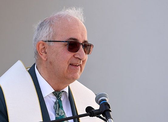 Ein älterer Mann mit weißem Haar und einer Brille steht lächelnd vor einem Mikrofon mit Ständer. Er ist in einen weißen Umhang mit gelbem Rand und einer schwarz-weißen Krawatte gekleidet.