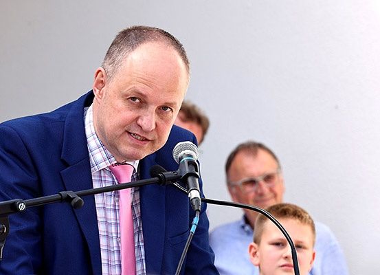 Ein Mann in einem blauen Anzug und einer pinken Krawatte spricht in ein Mikrofon, hinter ihm stehen zwei Personen, eine davon ist ein Junge.