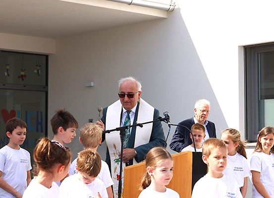 Ein älterer Mann steht mit einem Mikrofon an einem Rednerpult, trägt eine weiße Stola und spricht eine Gruppe von Kindern in weißen T-Shirts vor einem Gebäude an.