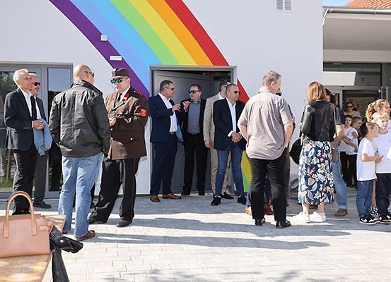 Eine Gruppe von Erwachsenen und Kindern steht vor einem Gebäude mit einer Regenbogenwand, sie schauen in das Gebäude hinein.