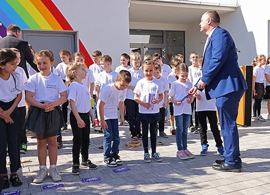 Eine Gruppe junger Kinder in weißen T-Shirts steht in einer Reihe vor einem Gebäude mit einem Regenbogen an der Wand, ein Mann in einem blauen Anzug spricht sie an.