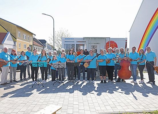 Eine Gruppe von Menschen in blauen Shirts, die Saiteninstrumente halten, posiert für ein Foto im Freien mit einem Regenbogen an der Wand und Gebäuden im Hintergrund.