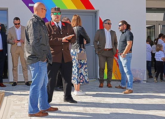 Eine Gruppe formell gekleideter Männer und Frauen steht vor einem Gebäude mit einem Regenbogen-Mural. Ein Mann trägt eine Militäruniform und grüne Sonnenbrille.