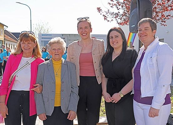 Fünf Frauen stehen zusammen vor einem Regenbogenbanner. Die Frau in der Mitte trägt eine Halskette und eine Uhr. Hinter ihnen stehen Gebäude und eine Straßenlaterne.