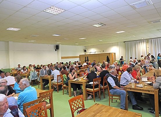 Viele Menschen sind in einem großen Raum versammelt, sitzen an Tischen und Stühlen. Einige essen und trinken, andere unterhalten sich. Der Raum hat einen grünen Boden und weiße Wände.