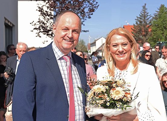 Ein Mann und eine Frau lächeln für ein Foto. Die Frau hält einen Blumenstrauß. Sie stehen draußen, mit Leuten hinter ihnen.