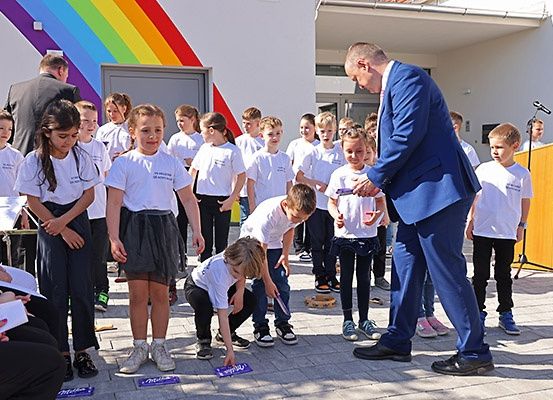 Eine Gruppe von Kindern in weißen T-Shirts ist um einen Mann in einem blauen Anzug versammelt. Er kniet und verteilt Karten an die Kinder. Die Kinder schauen zu ihm und einige halten Musikinstrumente. Hinter ihnen steht ein Gebäude mit einer regenbogenfarbenen Wand.