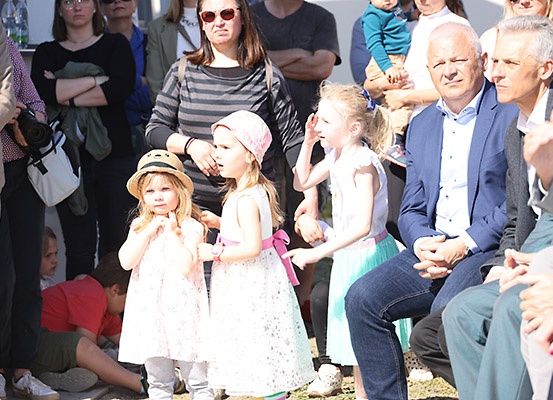Eine Gruppe von Menschen, einschließlich Erwachsener und Kindern, versammelt sich im Freien. Drei junge Mädchen, eines mit rosa Hut und die anderen in weißen Kleidern, stehen vor einem sitzenden Publikum. Erwachsene umgeben die Kinder, einige stehend und andere sitzend.