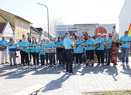 Eine Gruppe von Menschen in blauen Hemden und schwarzen Hosen steht in einem Außenbereich, hält Musikinstrumente und führt vielleicht eine Aufführung durch. Sie befinden sich vor einem Gebäude mit weißer Wand und einem Regenbogen.