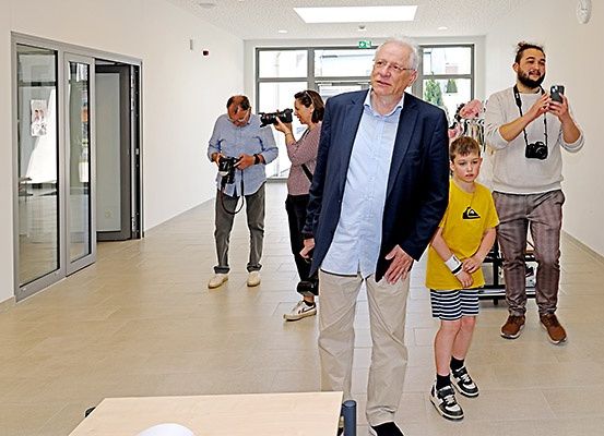 Ein Mann im Anzug führt eine Gruppe von Menschen durch einen Raum. Zwei Männer mit Kameras fotografieren ihn.