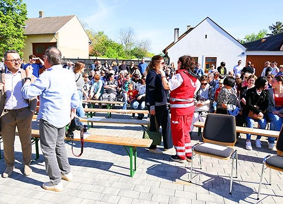 Eine Menschenmenge versammelt sich in einem Außenbereich mit Bänken. Einige stehen, andere sitzen. Eine Person trägt ein rotes Uniform. Im Hintergrund befinden sich Gebäude und Bäume.