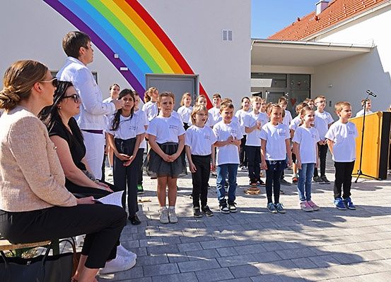 Kinder in weißen Shirts stehen in einer Reihe, möglicherweise für eine Schulveranstaltung, mit einem Regenbogen an der Wand hinter ihnen.