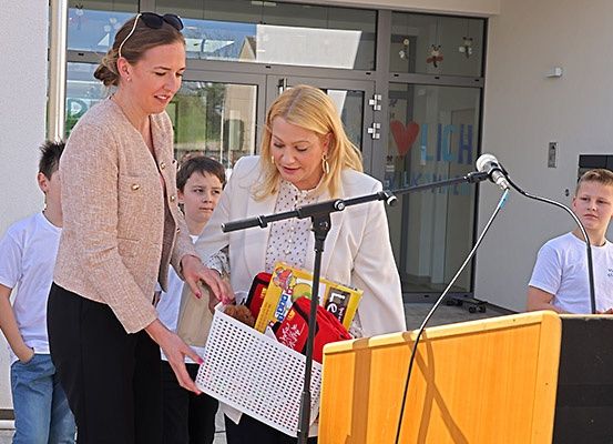 Zwei Frauen, eine präsentiert der anderen einen Korb, stehen an einem Podium mit einem Mikrofon davor. Dahinter steht eine Gruppe von Kindern.