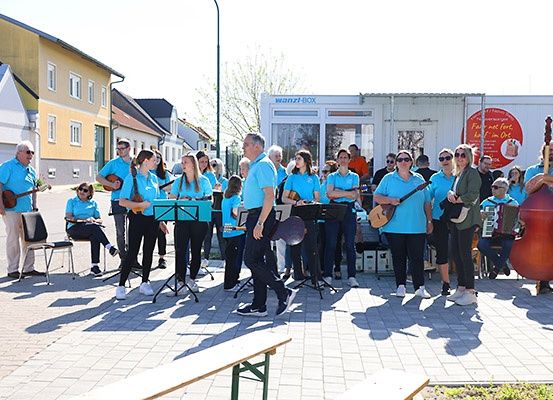 Eine Gruppe von Menschen in blauen T-Shirts spielt Musikinstrumente im Freien. Sie stehen auf einem gepflasterten Weg. Einige von ihnen halten Gitarren, während andere Akkordeons und Celli spielen. Dahinter befindet sich ein weißes Gebäude mit einer Glastür und einem roten Schild.