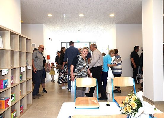 Mehrere Menschen stehen und unterhalten sich in einem Flur mit weißen Wänden und gefliestem Boden. Ein Regal mit Büchern steht links. Ein Tisch mit Blumen und Stühlen steht im Vordergrund.
