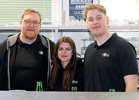 Drei Personen in schwarzen Shirts posieren für ein Foto vor einem Tresen mit grünen Flaschen. Dahinter steht ein Regal mit Gläsern, und ein Plakat bewirbt eine kroatische Nachtveranstaltung.