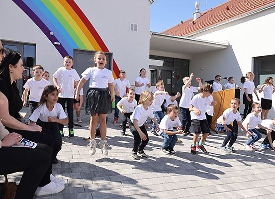 Eine Gruppe von Kindern in weißen Hemden und schwarzen Röcken führt vor einem Gebäude mit Regenbogendesign auf. Zwei Frauen sitzen und schauen zu.