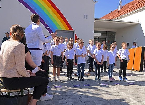 Eine Gruppe von Kindern in weißen T-Shirts steht in einer Reihe und singt und klatscht. Ein Erwachsener führt sie an, während ein anderer Erwachsener von einem Sitzplatz aus zusieht. Ein Gebäude mit Regenbogenhintergrund und einem Lautsprecher steht hinter ihnen.