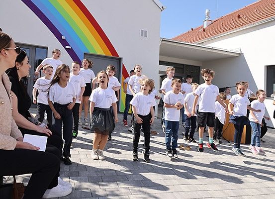 Kinder in weißen T-Shirts singen vor einem Gebäude mit einem Regenbogenwand. Eine erwachsene Frau sitzt neben ihnen.