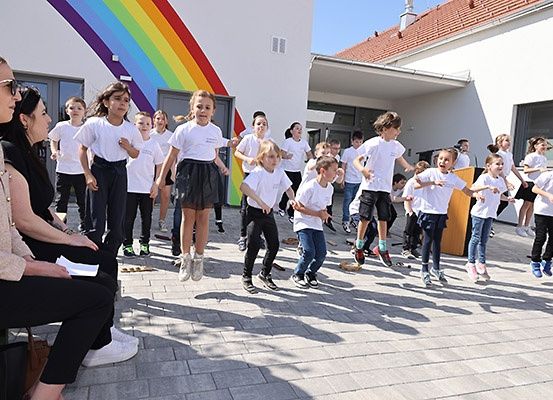 Eine Gruppe von Kindern in weißen T-Shirts und schwarzen Hosen springt und tanzt vor einem Gebäude mit einem Regenbogen an der Wand.