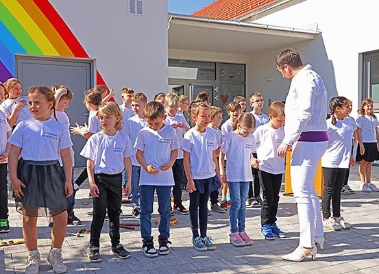 Eine Gruppe von Kindern in weißen T-Shirts steht vor einem Gebäude, mit einem Mann vor ihnen. Das Gebäude hat einen Regenbogen an der Wand gemalt.