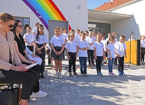 Eine Gruppe von Kindern in weißen T-Shirts steht vor einem Gebäude mit einem Regenbogen-Mural. Zwei Frauen sitzen vor ihnen.
