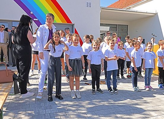 Eine Gruppe von Kindern und Erwachsenen steht vor einem Gebäude mit einem Regenbogen an der Wand. Einige Kinder tragen weiße Hemden.