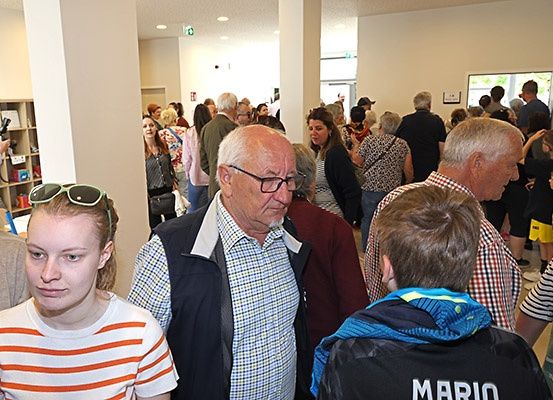 Eine Gruppe von Menschen ist drinnen, einige mit Brille, stehen und unterhalten sich. Ein Mann in einem karierten Hemd schaut nach vorne. Ein anderer Mann mit einer Sonnenbrille auf dem Kopf ist in der Nähe.