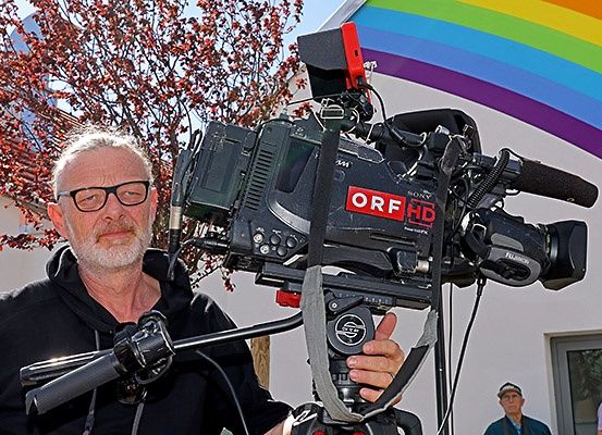Ein Mann mit Brille hält eine professionelle Kamera mit der Aufschrift ORF HD vor einem Gebäude mit Regenbogenhintergrund.