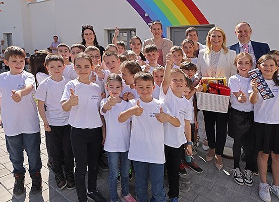 Gruppenfoto von Kindern und Erwachsenen in weißen T-Shirts, einige Erwachsene halten Geschenke, posieren vor einem Gebäude mit Regenbogenhintergrund.