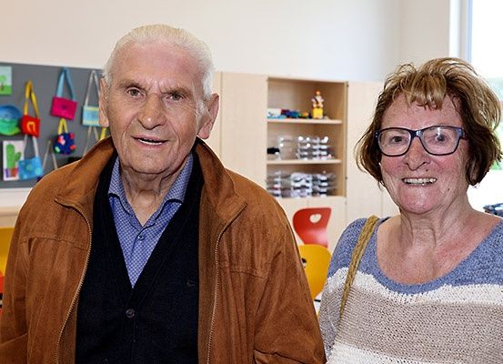 Zwei Personen stehen in einem Raum mit Regalen, bunten Stühlen und einer Pinnwand mit Zeichnungen. Ein Mann trägt eine braune Jacke und eine Frau trägt eine Brille.