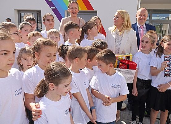 Eine Gruppe von Kindern in weißen T-Shirts steht zusammen vor einem Gebäude, vor dem zwei Frauen und ein Mann lächelnd auf sie schauen.