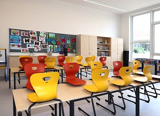 Ein Klassenzimmer mit bunten Stühlen und Tischen. Die Wände sind weiß, und es gibt eine Pinnwand mit verschiedenen Kunstwerken. Der Raum hat Glasfenster mit Blick auf einen grünen Baufahrzeug.