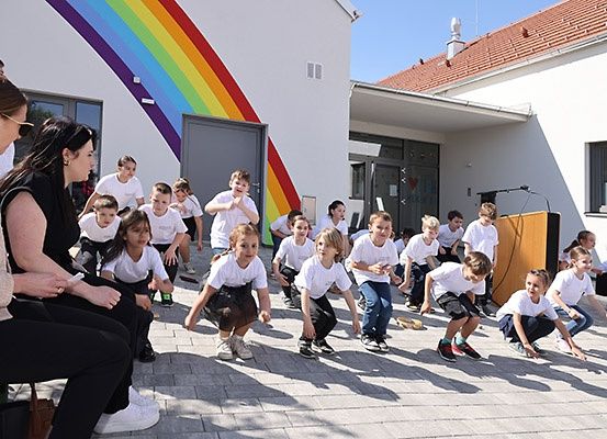 Mehrere Kinder in weißen Hemden machen Ausfallschritte auf einem Bürgersteig vor einem Gebäude mit einem Regenbogen an der Wand.